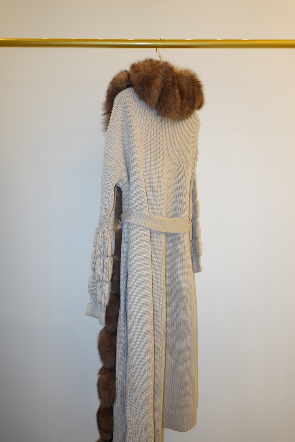 The back of a brown fur-trimmed jackets/cardigan on a rack against a white wall.